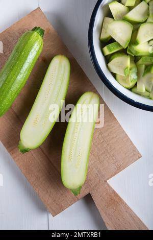 One green zucchini with two section halves on a cutting board with chopped zucchini in a bowl on a wooden background. Stock Photo