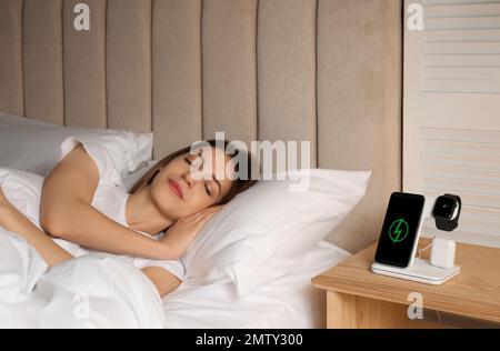 Smartphone, watch, earphones charging on wireless pad and sleeping woman in room Stock Photo