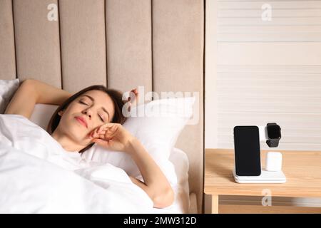 Smartphone, watch, earphones charging on wireless pad and sleeping woman in room Stock Photo