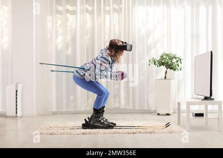 Full length profile shot of a young woman skiing and wearing a virtual reality headset in front of tv at home Stock Photo