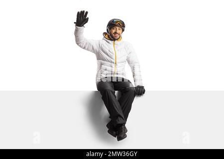 Young man wearing a skiing equipment sitting on a blank panel and waving isolated on white background Stock Photo