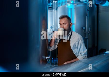 Brewer tasting fresh brewed beer while standing in front of metal containers Stock Photo