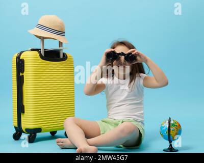 Curious little traveler child girl looking at camera through vintage binoculars, sitting near yellow suitcase and globe, isolated over blue color back Stock Photo