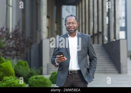 Portrait of a successful African American male attorney, lawyer. Who is standing in glasses and a suit outside near the courthouse, holding a phone in his hand, smiling at the camera. Stock Photo