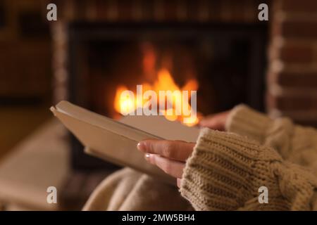 Woman reading book near burning fireplace at home, closeup Stock Photo