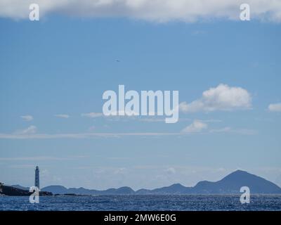 a view of Guadeloupe lighthouse on rocky outcrop on left, framed at top by cloud and bottom by sea and silhouette of coastline in a blue sky Stock Photo