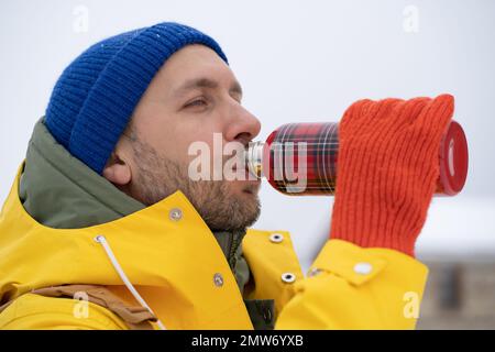 European unshaved man dressed blue beanie, orange knitted gloves drinks hot tea from therm bottle. Stock Photo