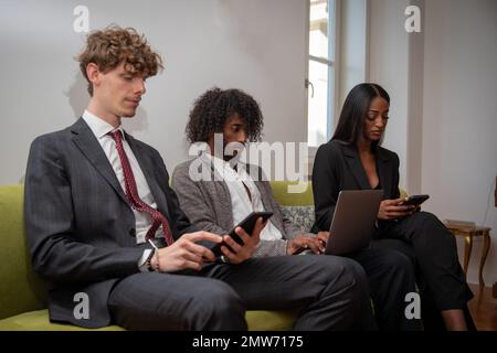 Three colleagues use their tech devices while sitting on the sofa at their workplace Stock Photo