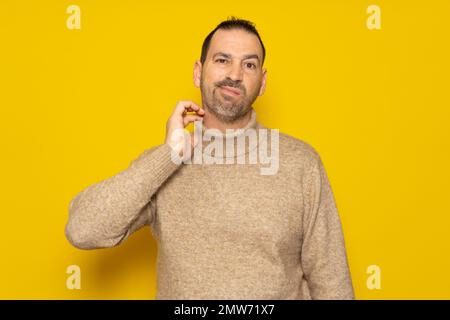 Bearded hispanic man wearing beige turtleneck scratching his neck and grimacing isolated over yellow background. Concept of doubt and indecision. Stock Photo