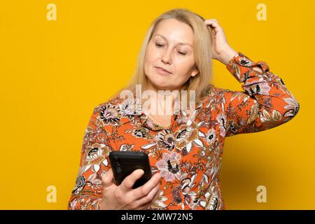 What to text. Portrait of pensive woman holding mobile phone and looking away scratching head with hand isolated over yellow studio background Stock Photo
