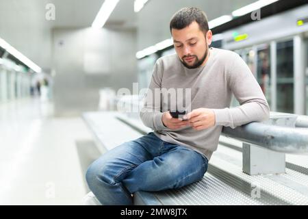 Bearded man sitting with phone on bench on subway platform Stock Photo