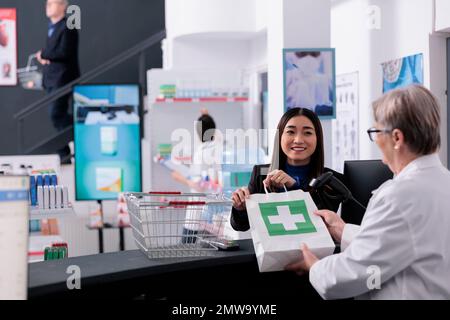 Asian woman buying supplements at pharmacy store counter desk, taking medicaments package from pharmaceutical assistant. Happy young customer getting purchase at cash register Stock Photo