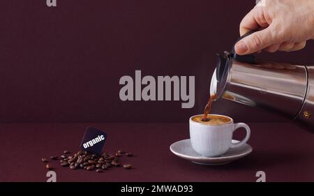 A hand pouring coffee from a mocha pot into a small cup next to a pile of coffee beans against a dark burgundy background. Stock Photo