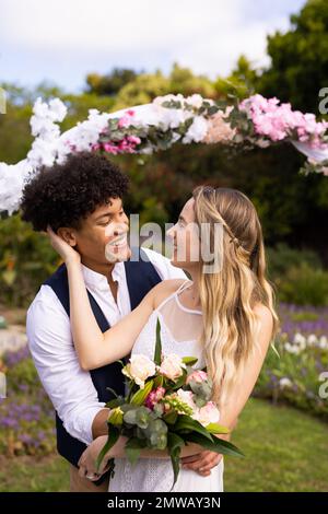 Vertical of happy diverse bride and groom holding bouquet embracing at outdoor wedding Stock Photo