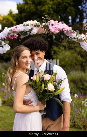 Vertical portrait of smiling diverse bride and groom embracing at outdoor wedding Stock Photo