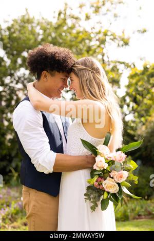 Vertical of happy diverse bride and groom with bouquet embracing at wedding in garden Stock Photo