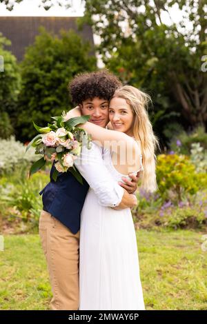 Vertical portrait of happy diverse bride and groom embracing at wedding in garden Stock Photo