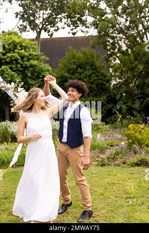 Vertical of happy diverse bride and groom holding hands, dancing at wedding in garden, copy space Stock Photo