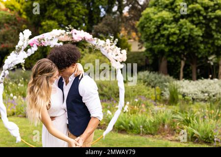 Happy diverse bride and groom holding hands, dancing at wedding in garden, with copy space Stock Photo