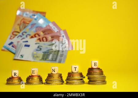 German word for pension, Rente, from wood cubes on stacks of coins. Euro cash money on yellow background, coins and banknotes, close up. Stock Photo