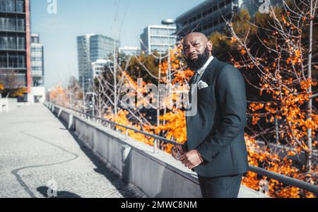 A balding black man with a full unshaven beard, formally dressed in a dark suit and polka-dot tie, holding his reading glasses, on the sidewalk in an Stock Photo