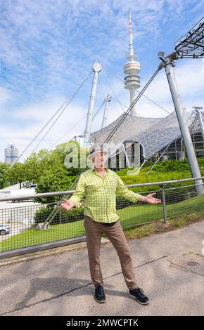 Friendly smiling man at the Olympic tower with Olympic tent roof, BMW tower on the left, Olympic Park, Olympic grounds, Munich, Upper Bavaria Stock Photo
