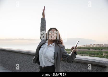 A businesswoman smiles with one arm raised and her tablet in hand. Freedom and remote work concept Stock Photo