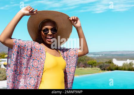 Portrait of happy african american woman wearing straw hat and sunglasses by swimming pool Stock Photo