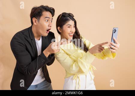 Shocked excited Asian businessman along with happy smiling woman pointing finger at smart phone over beige studio background. Man in white shirt and suit and woman in yellow top and white trousers. Stock Photo