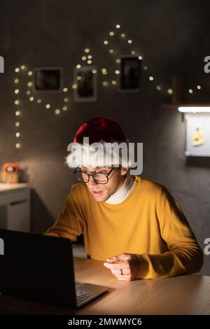 Portrait of young man working on a laptop while sitting at his desk at dormitory. In the evening creative man in santa hat works on a computer in cozy Stock Photo