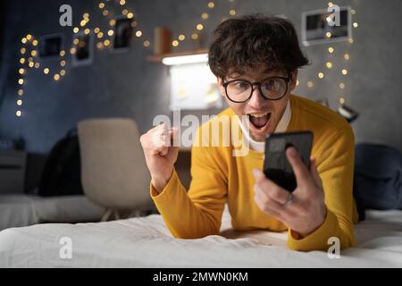 Excited happy young arabic man winner feeling joy using smartphone winning lottery game, betting, getting cashback online gift holding cell phone lyin Stock Photo