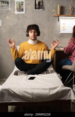 Young male university student meditating in lotus position on bed relaxing in dorm room. Copy space Stock Photo