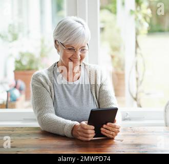 Browsing through family photos. A happy senior woman sitting at her kitchen table using a digital tablet. Stock Photo