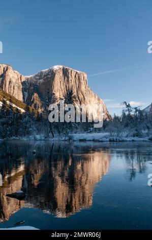The rocky surface of the Sierra Nevada mountains, reflecting early morning sunlight, being reflected in Merced river, on an early winter, snow-covered landscape in Yosemite national park. Stock Photo