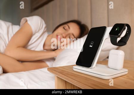 Smartphone, watch, earphones charging on wireless pad and sleeping woman in room Stock Photo