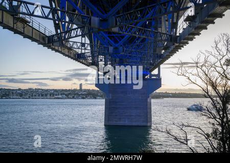Concrete pillars and steel beams under the Auckland Harbour Bridge illuminated in blue light. A motion blur ferry boat travelling towards the bridge. Stock Photo