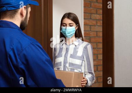 Young woman wearing medical mask receiving parcel from delivery man indoors. Prevention of virus spread Stock Photo