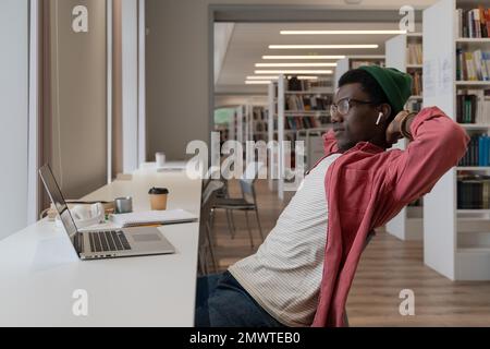 Relaxed Black student man sitting at desk with laptop in university library, looks educational video Stock Photo