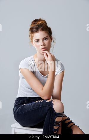 Confidence is the sexiest thing a woman can wear. Studio portrait of a beautiful young woman sitting on a stool against a gray background. Stock Photo