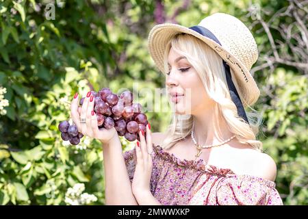 Photo of a young blonde woman in a hat, who is holding a bunch of grapes in her hand. Outdoor recreation,summer picnic. Stock Photo