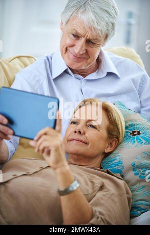Its so much easier to stay connected to our family. a happy elderly couple watching something on a digital tablet while relaxing on their sofa. Stock Photo