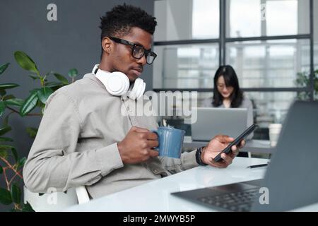 Serious African American businessman drinking coffee while reading message on smartphone at his workplace in modern office Stock Photo
