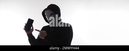 Hacker Anonymous and face mask with smartphone in hand. Man in black hood shirt holding and using mobile phone on white background. Represent cyber cr Stock Photo