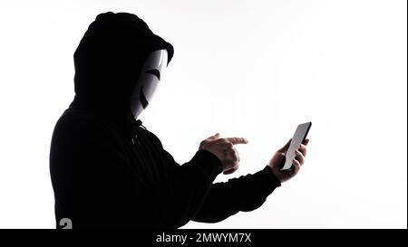 Hacker Anonymous and face mask with smartphone in hand. Man in black hood shirt holding and using mobile phone on white background. Represent cyber cr Stock Photo