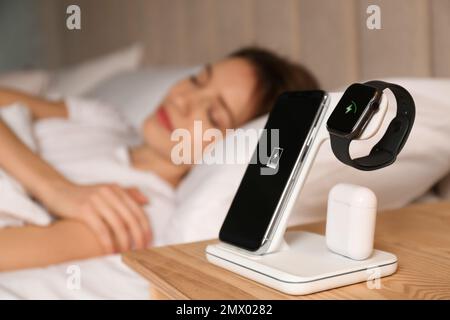 Smartphone, watch, earphones charging on wireless pad and sleeping woman in room Stock Photo