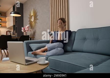 Young woman in the living room covering face with hand, scared to look at message at her cell phone Stock Photo