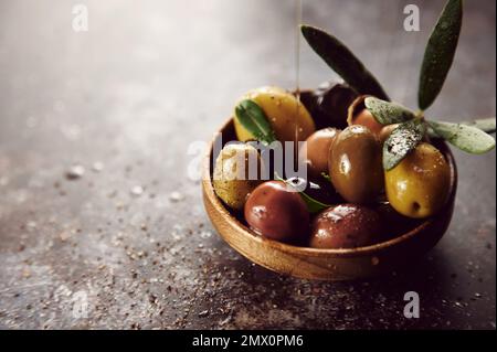 Olive oil jet pouring on mix of tasty marinated olives in bowl placed on gray background with seasoning in light kitchen Stock Photo