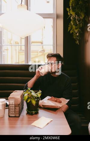 Man drinking coffee in a paper cup and having breakfast inside a coffee shop Stock Photo