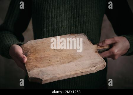 Crop unrecognizable female in warm knitted sweater holding worn out wooden cutting board in hands Stock Photo