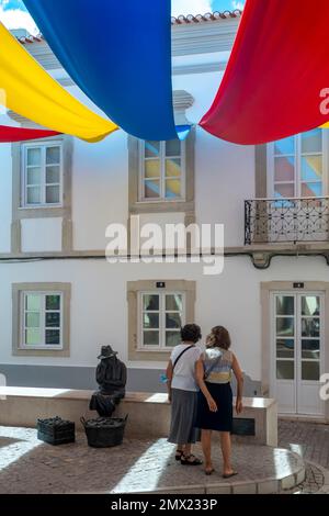 Two women watch a statue over a protection cloth in the street for creating shade in the city of Loule, near the market, in the Algarve, Portugal. Stock Photo